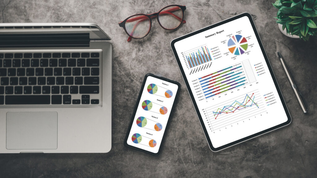 A sleek workspace showcasing a laptop, tablet with summary report, smartphone with charts, alongside eyeglasses and a green plant.